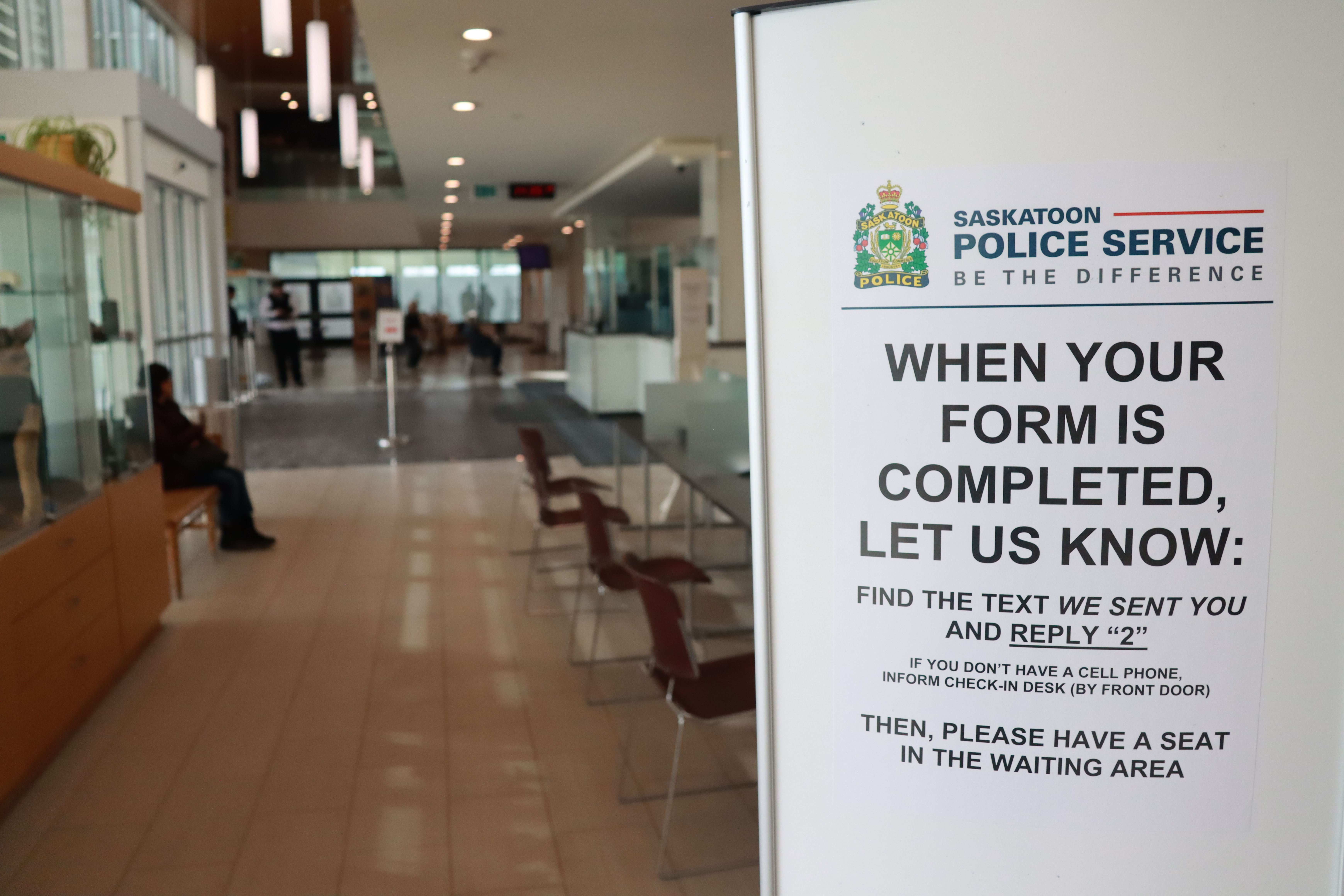 A photo of the Saskatoon Police Service front lobby. In the foreground on the right is a sign reading, 'When your form is completed, let us know: find the text we sent you and reply 2. if you don't have a cell phone, inform the check-in desk (by the front door). Then, please have a seat in the waiting area.' In the background of the photo are chairs next to a table on the right, and a person sitting on a bench next to a display case on the left.