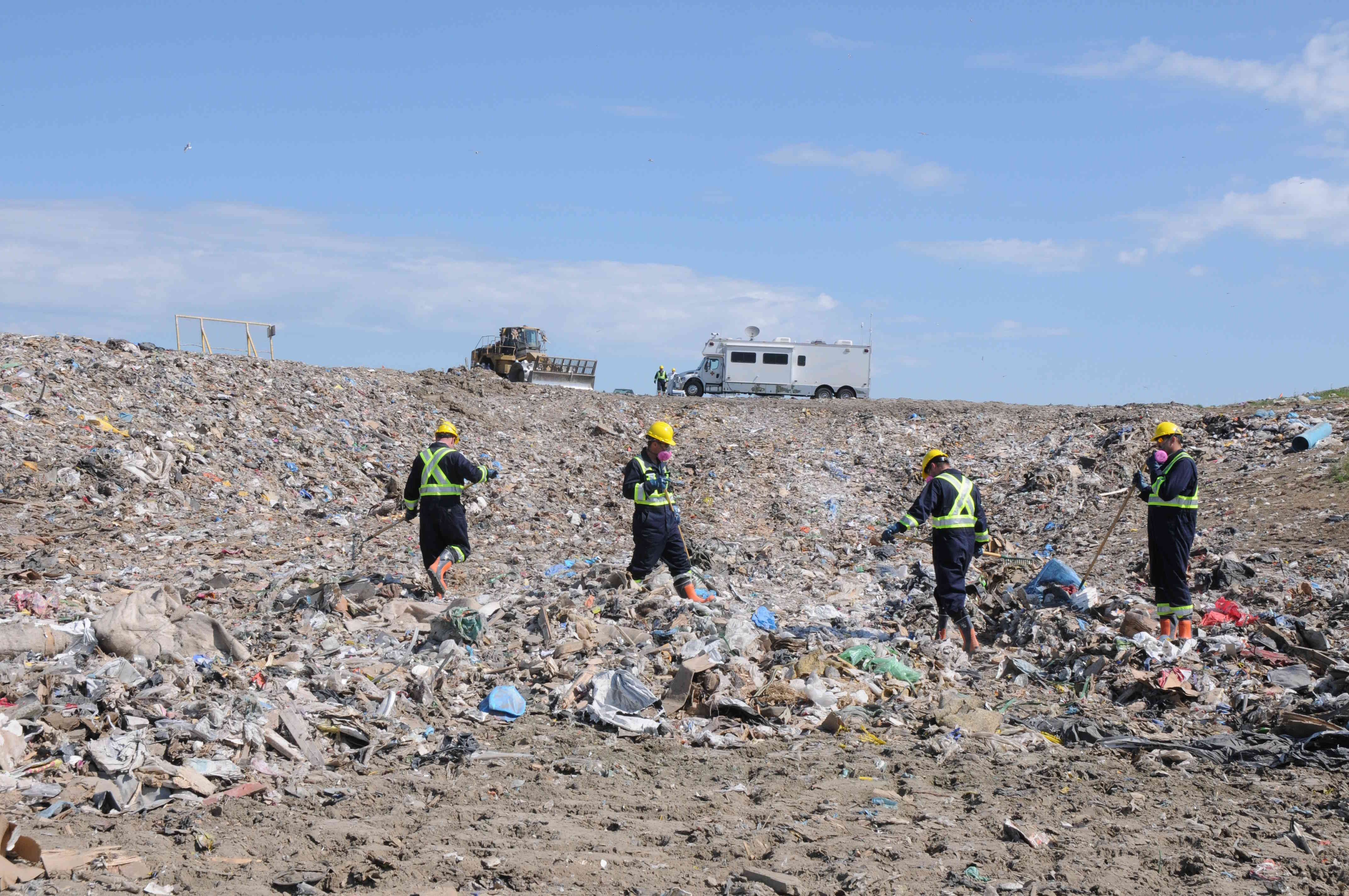 The command post and a loader are pictured in the background, with four workers searching through a landfill.