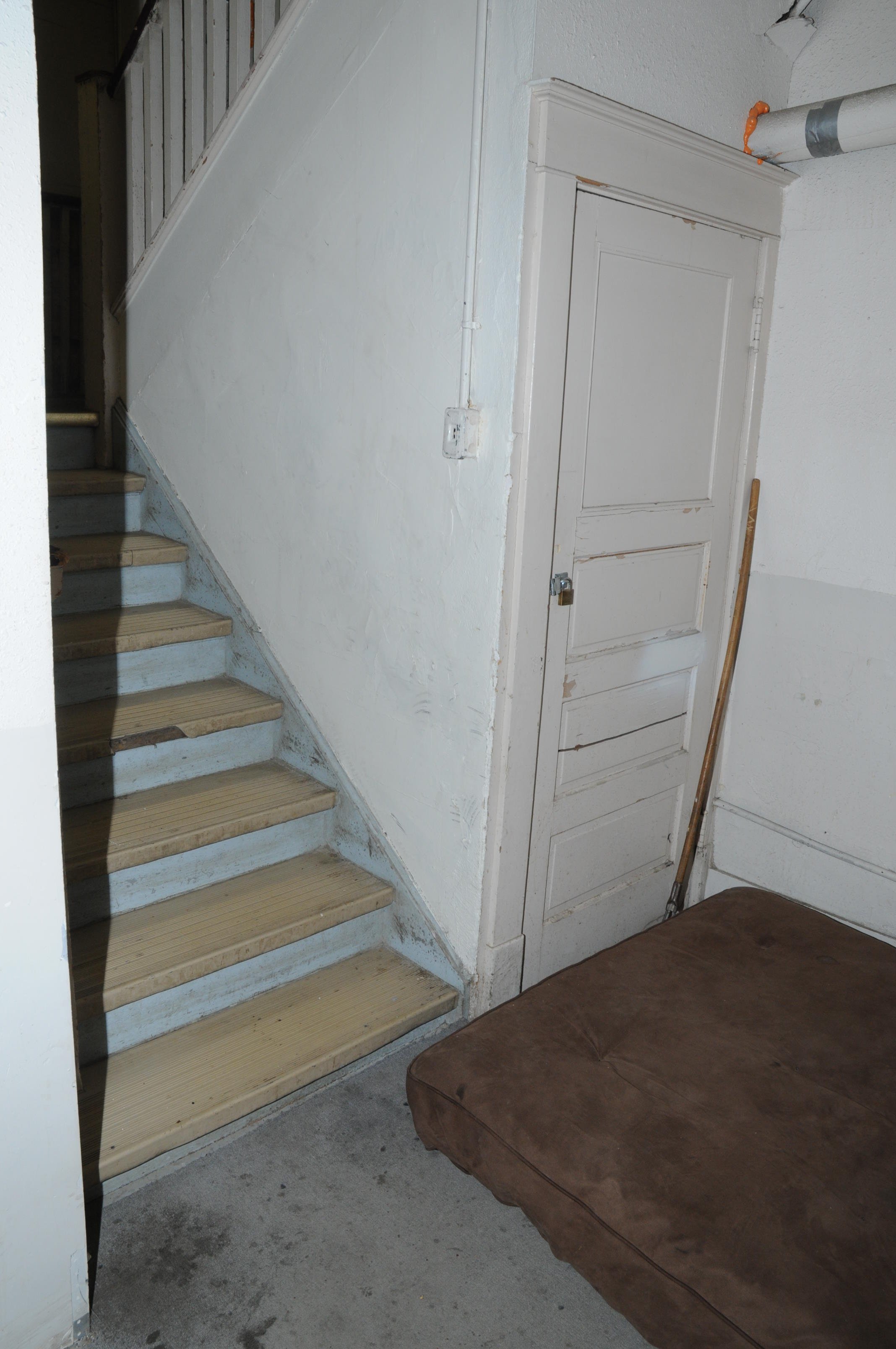 Single closed room at the bottom of a stairwell and a brown mattress on the floor, door is locked with a key lock.