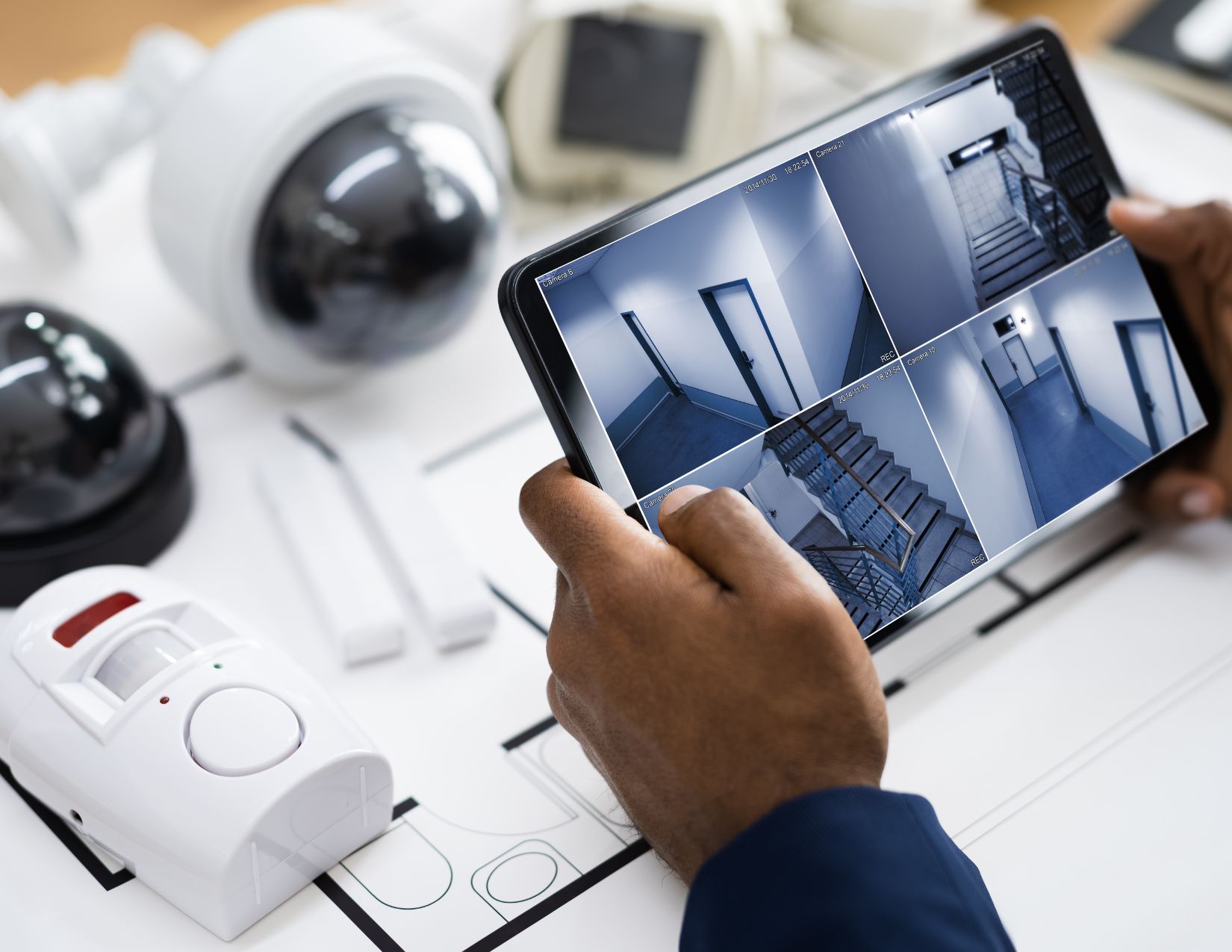 Two hands holding a tablet device. On the device's screen are four different camera angles; two displaying different stairwells, and two displaying different sets of doors. There are several different types of security cameras in the background.